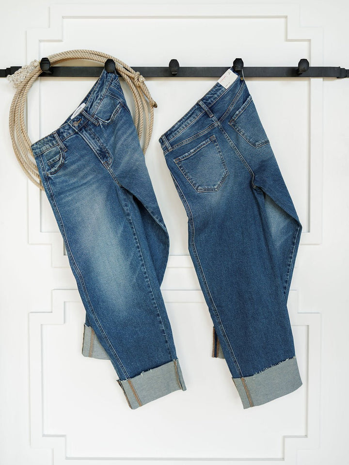 Two pairs of The Kamden High Rise Barrel Jeans by Savannah Sevens western life{&}style, in medium wash with rolled cuffs, hang on a black wall-mounted rack with a beige rope draped over it against a white paneled background.
