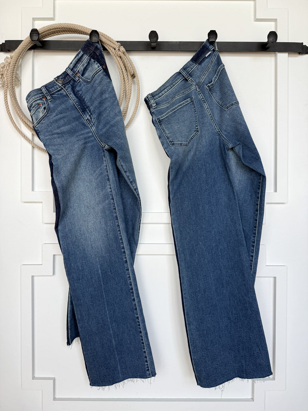 Two pairs of the Kanen High Rise Wide Leg Jean from Savannah Sevens western life{&}style, in a medium wash denim blue, hang side by side on black hooks against a white wall; a coiled rope is draped over the left hook.