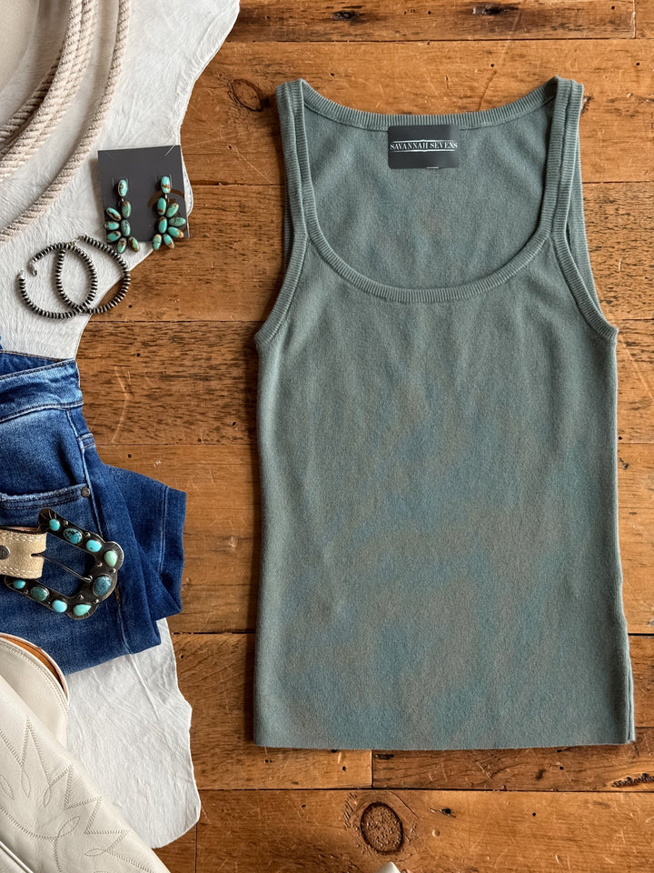 The Kaplan Blue Sage Scoop Tank by Savannah Sevens western life{&}style is displayed on a wooden surface, accompanied by denim jeans, turquoise jewelry, a white hat with a rope band, and white cowboy boots.