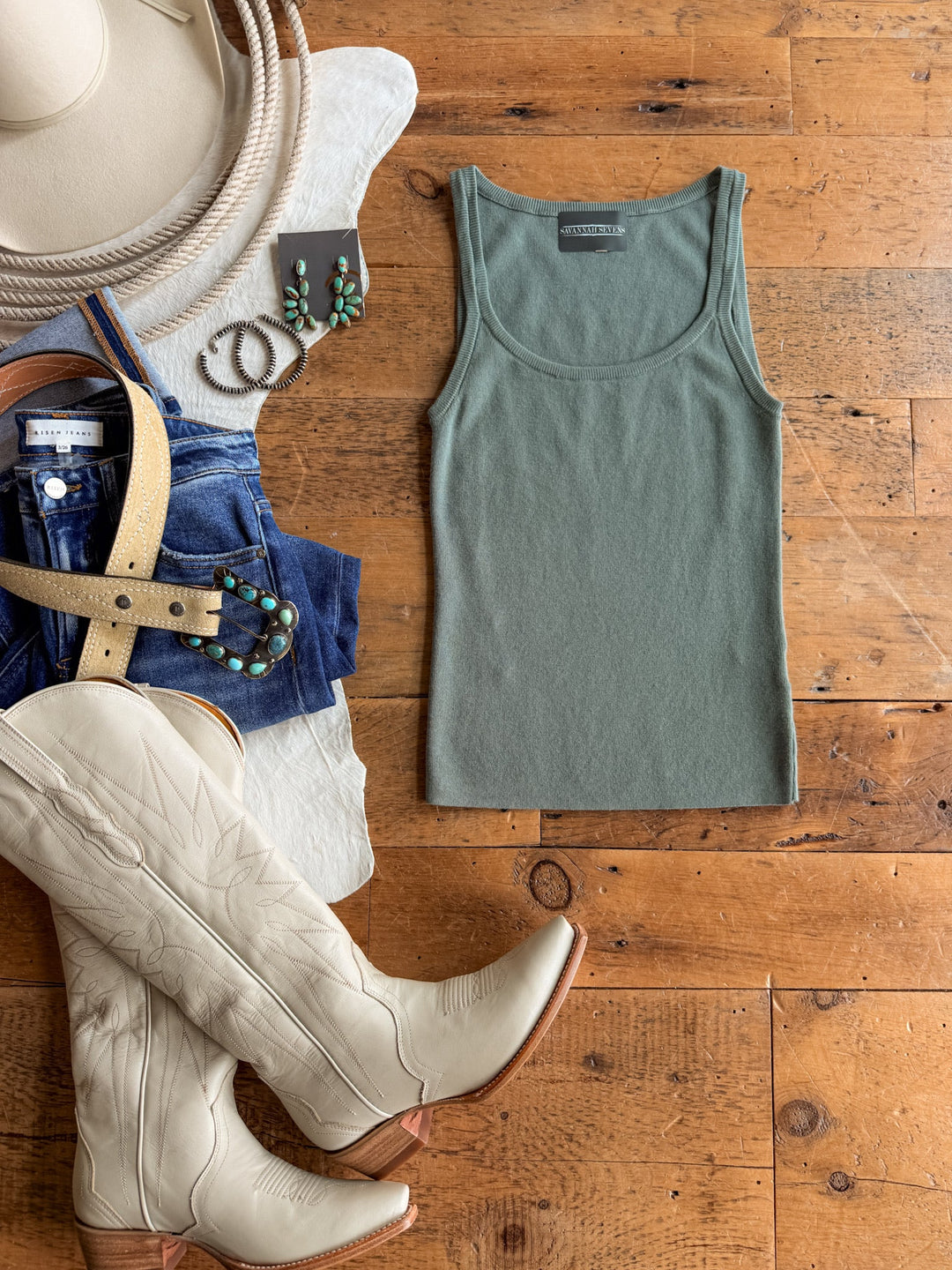 The Kaplan Blue Sage Scoop Tank by Savannah Sevens western life{&}style is styled in a flat lay on a wooden floor, paired with cowboy boots, jeans, a beige hat with rope detail, a belt, stud earrings, and turquoise jewelry to create a western-inspired outfit.