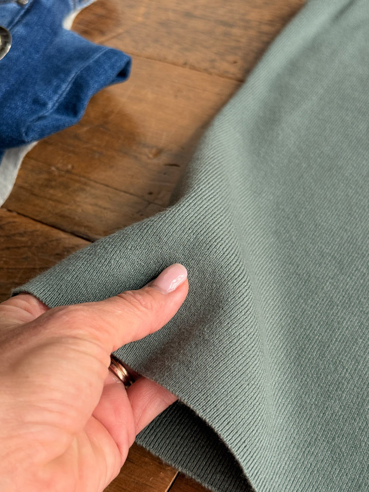 A hand with light pink nail polish holds the edge of a soft, green knit fabric on a wooden surface with the Kaplan Blue Sage Scoop Tank by Savannah Sevens western life{&}style nearby.
