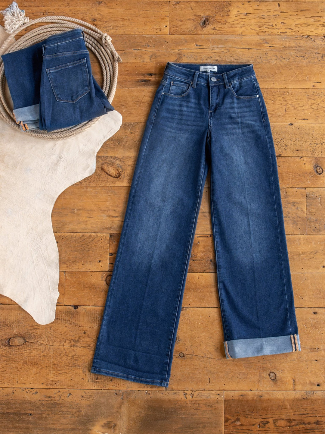 The Kase High Rise Wide Leg Jean by Savannah Sevens western life{&}style is laid flat on a wooden floor, beside a folded pair of jeans, a coiled rope, and a light-colored animal hide rug.