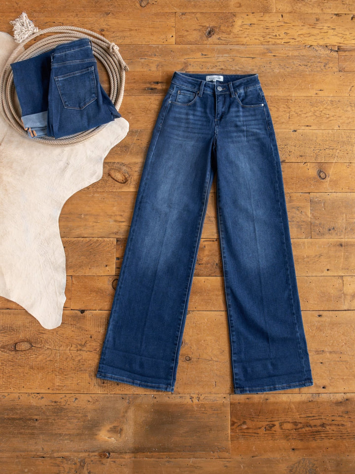 The Kase High Rise Wide Leg Jean by Savannah Sevens western life{&}style is laid flat on a wooden floor, with another pair of jeans folded in a circular woven basket nearby atop a light cowhide rug.