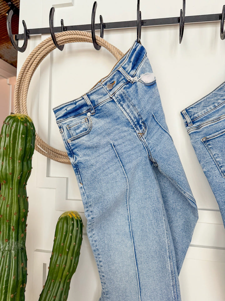 The Kasyn High Rise Slim Wide Leg Jean by Savannah Sevens western life{&}style in light blue hangs from black hooks on a white wall, flanked by two tall green cacti. Another pair of vintage wash jeans is partially visible on the right.