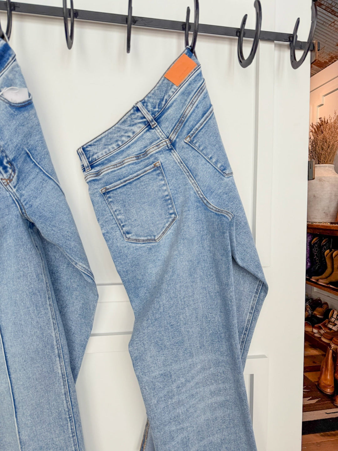 A pair of The Kasyn High Rise Slim Wide Leg Jeans by Savannah Sevens western life{&}style hangs on a black hook rack over a white paneled door, with another pair partly visible and shelves with shoes, boots, and décor in the background.