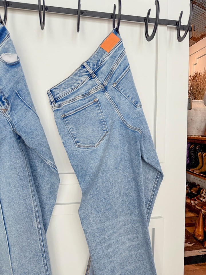 A pair of The Kasyn High Rise Slim Wide Leg Jeans by Savannah Sevens western life{&}style hangs on a black hook rack over a white paneled door, with another pair partly visible and shelves with shoes, boots, and décor in the background.