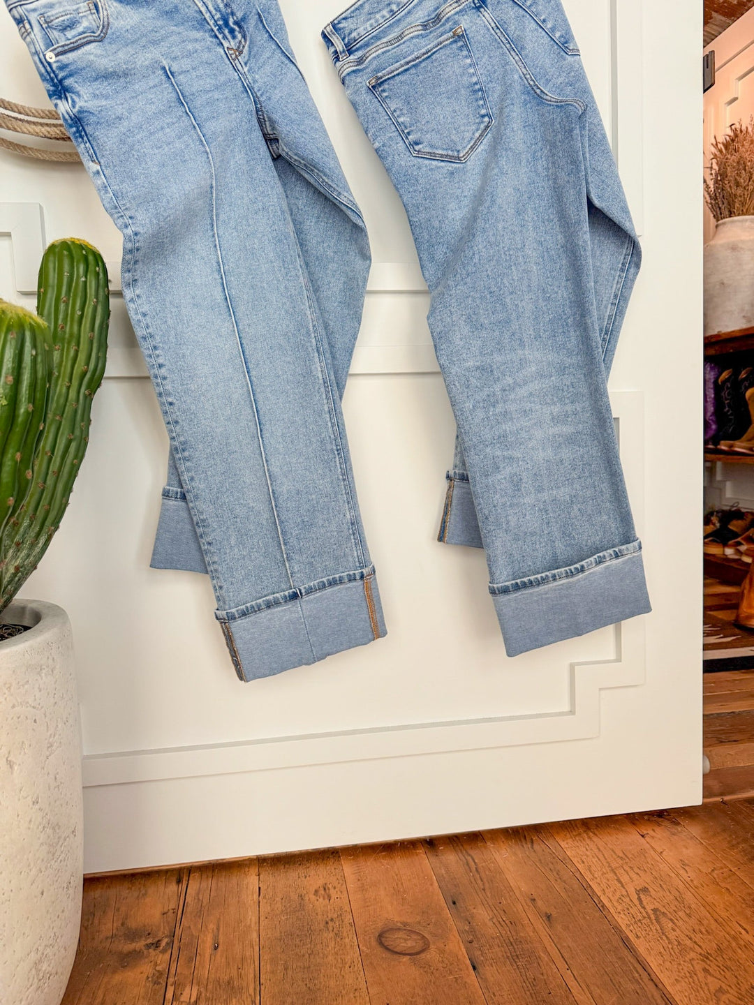 Two pairs of The Kasyn High Rise Slim Wide Leg Jeans by Savannah Sevens western life{&}style hang against a white wall, with a green potted cactus on the left and a wooden floor visible at the bottom.