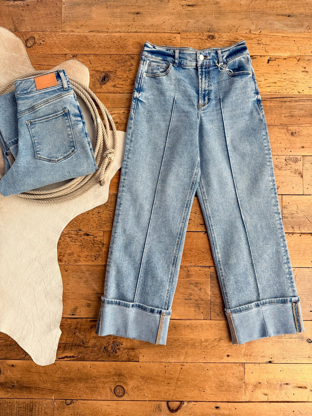 The Kasyn High Rise Slim Wide Leg Jean by Savannah Sevens western life{&}style is laid flat on a wooden floor, while a folded Dear John Denim pair with leather patch rests on a cowhide rug beside coiled rope.