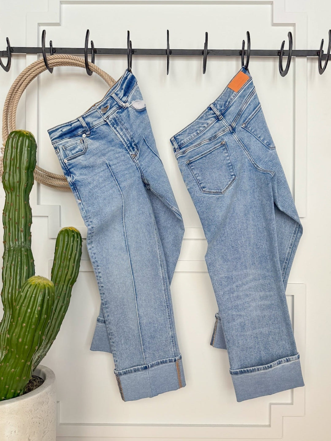 Two pairs of Savannah Sevens western life{&}style The Kasyn High Rise Slim Wide Leg Jeans in light blue hang from black hooks on a white panel. A tall green cactus in a white pot stands to the left.
