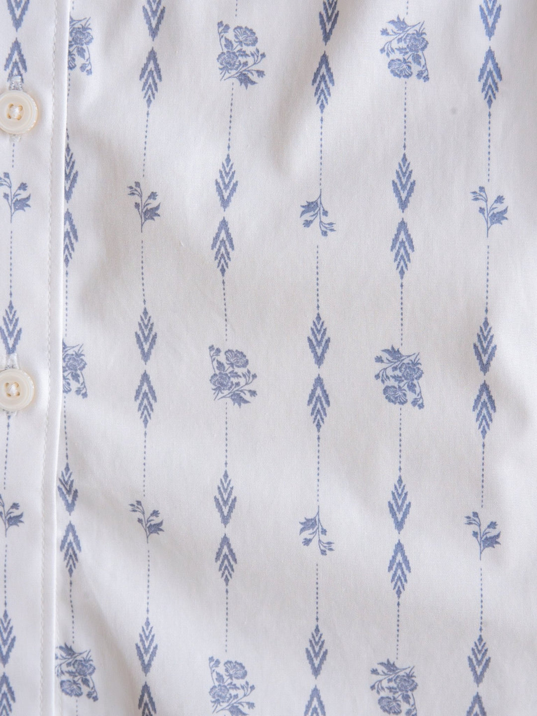 Close-up of the Kearney Ariat Southwest Floral Performance Shirt by Savannah Sevens western life{&}style in white, showcasing vertical blue arrows and small floral designs. Long sleeve button-up with white buttons is partially visible.