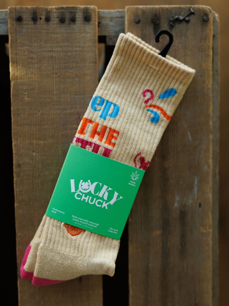 A pair of Keep The Faith Crew Socks by Savannah Sevens western life{&}style in a beige hemp blend with colorful designs, pink toes, and green Lucky Chuck label rests on rustic wood. Breathable socks with black hanger attached.