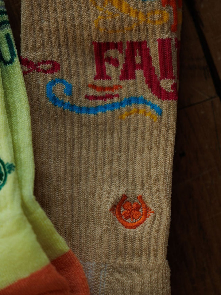 A close-up of the Savannah Sevens western life{&}style Keep The Faith Crew Socks in beige, featuring colorful text and a small embroidered orange horseshoe, beside a partially visible yellow-green sock on a wooden surface.