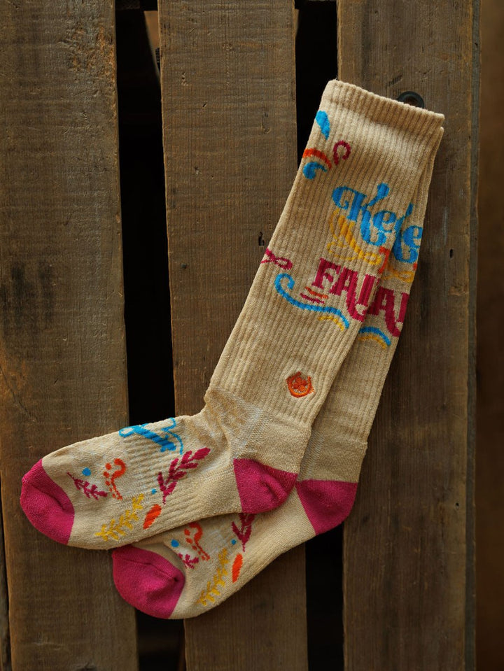 The Keep The Faith Crew Socks by Savannah Sevens western life{&}style are beige hemp blend with bold blue, red, and orange patterns and pink heels and toes, shown on a rustic wooden surface.