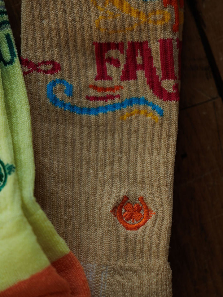 A close-up of the Savannah Sevens western life{&}style Keep The Faith Crew Socks in beige, featuring colorful text and a small embroidered orange horseshoe, beside a partially visible yellow-green sock on a wooden surface.