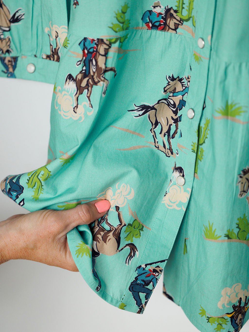 A hand with peach-colored nail polish holds the hem of the Kemah Seafoam Western Print Dress by Savannah Sevens western life{&}style, featuring a playful cowboy cactus print and white snap buttons for a fun, western-inspired look.