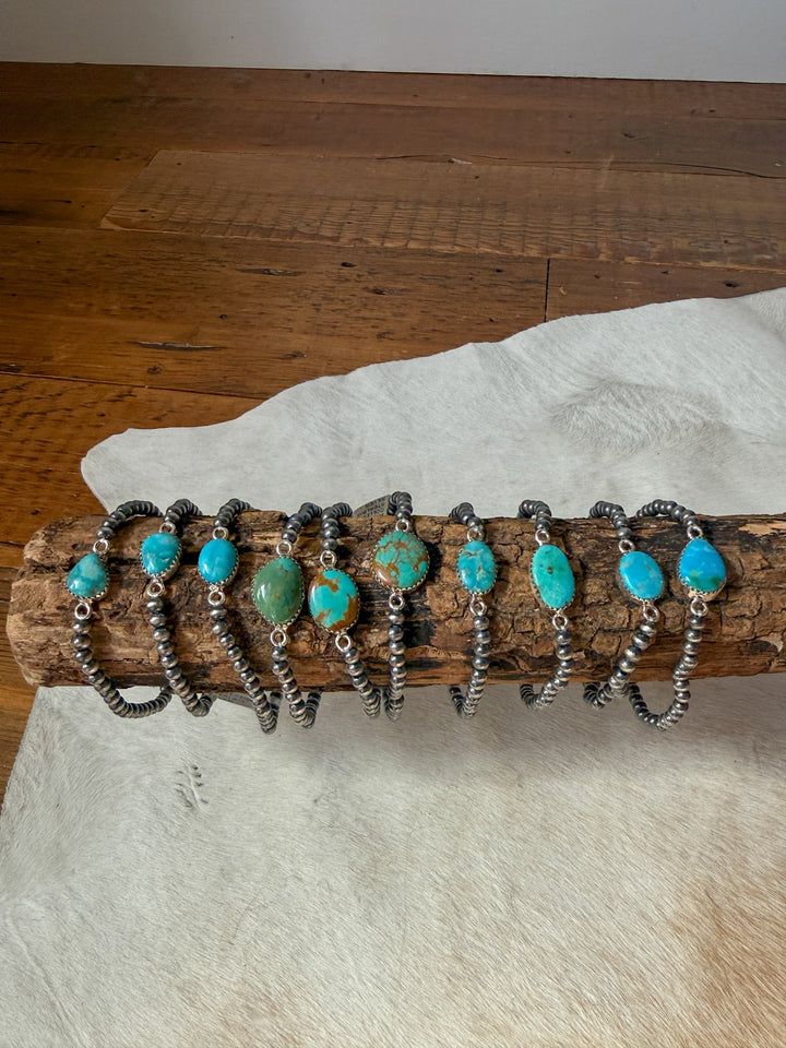 Nine Savannah Sevens western life&style Kemah Turquoise Stone Bracelets, each featuring sterling Navajo pearl beads and a single turquoise stone in various shapes and sizes, displayed in a row on a wooden log.