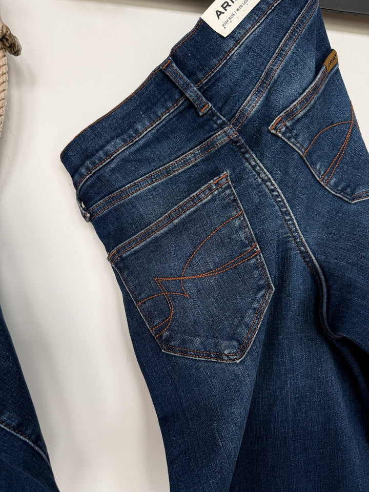 A close-up view of the back pocket and waistband of blue Kemp High Rise Wide Leg Jeans by Savannah Sevens western life{&}style, featuring orange stitching and a small brown label above the pocket, hanging on a white background.