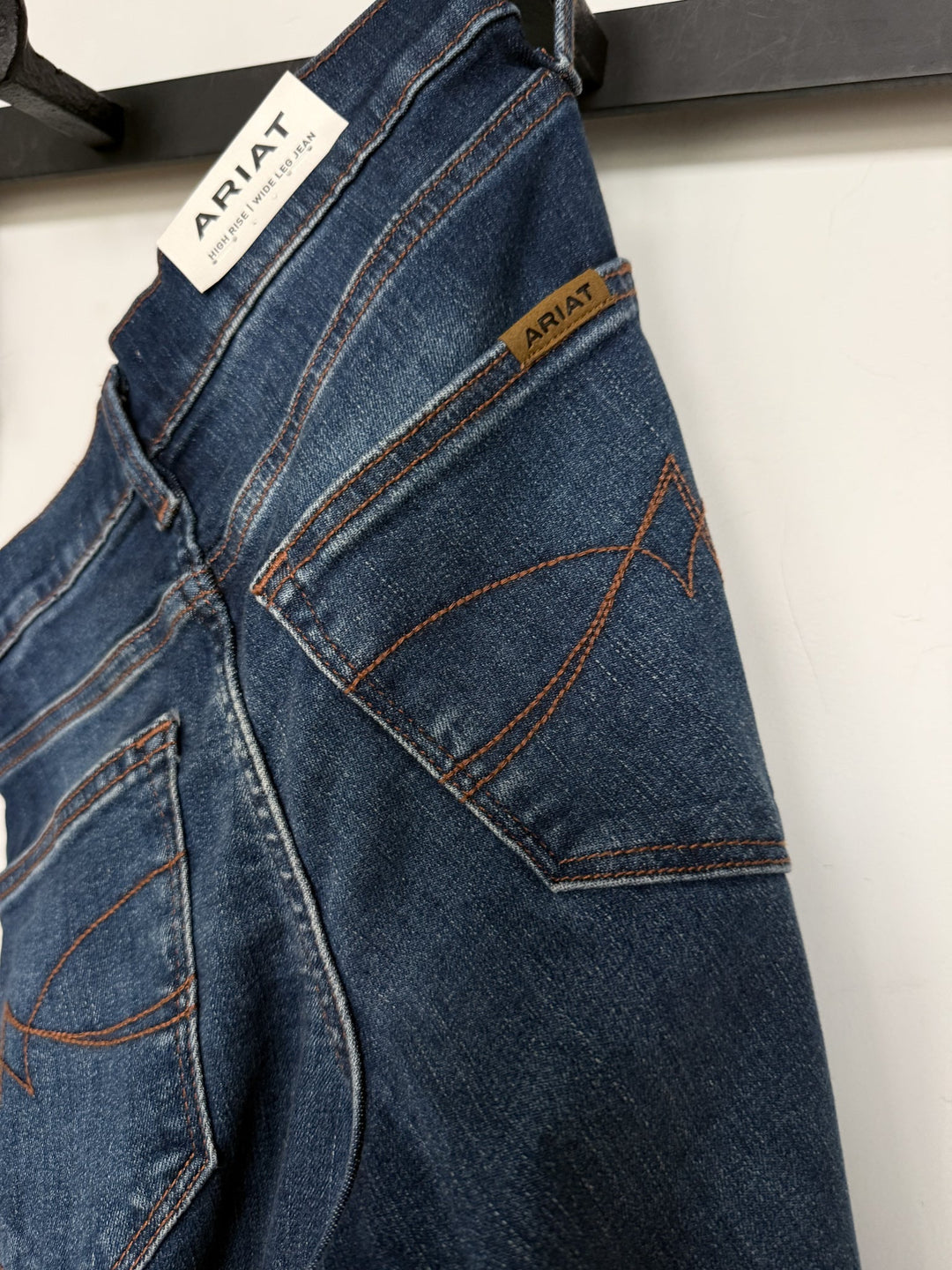 A pair of blue Kemp High Rise Wide Leg Jeans from Savannah Sevens western life{&}style hangs on a black hanger, displaying the back pocket with orange stitching and brand tags.