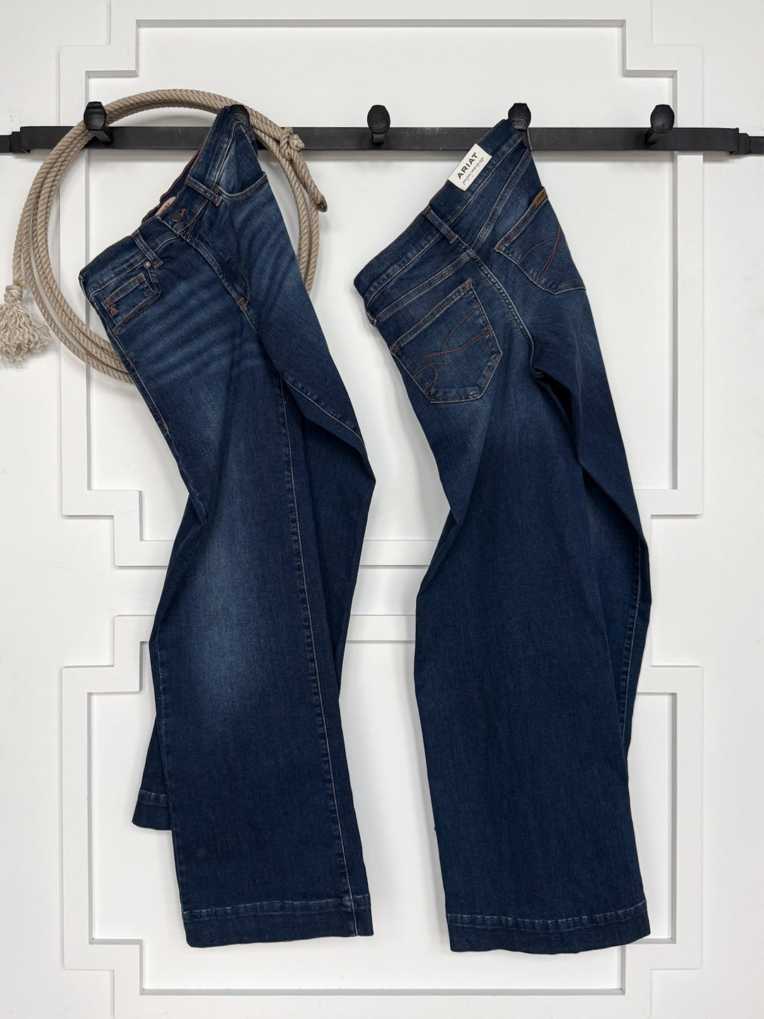 Two pairs of dark blue jeans, including the Kemp High Rise Wide Leg Jean by Savannah Sevens western life{&}style, hang on black hooks against a white wall, with a coiled beige rope draped over one hook.