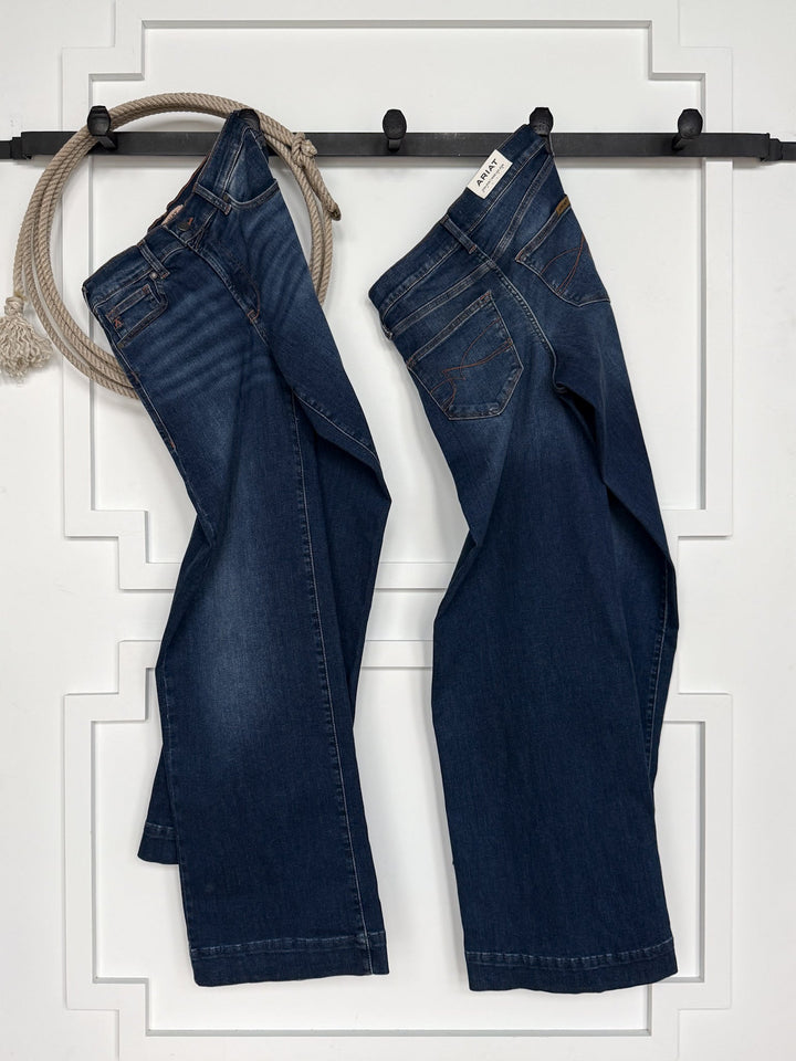 Two pairs of dark blue jeans, including the Kemp High Rise Wide Leg Jean by Savannah Sevens western life{&}style, hang on black hooks against a white wall, with a coiled beige rope draped over one hook.