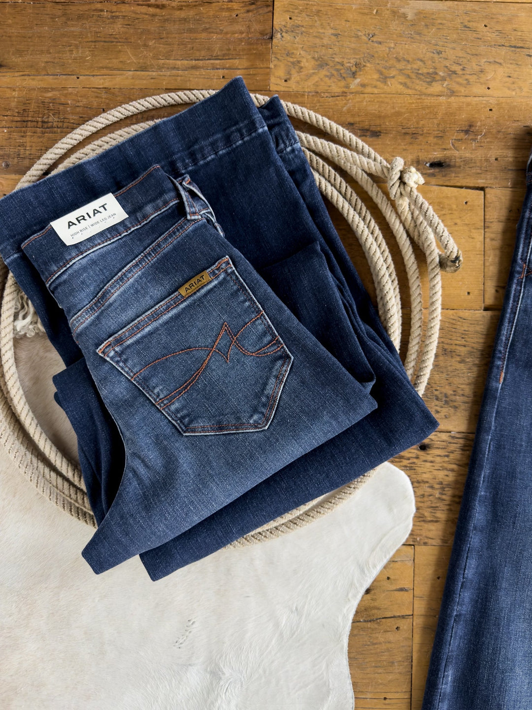 A folded pair of dark blue Kemp High Rise Wide Leg Jeans by Savannah Sevens western life{&}style rests on another pair of jeans atop a coiled rope on a wooden floor, next to a white cowhide rug. These jeans combine high-rise comfort with wide leg style.