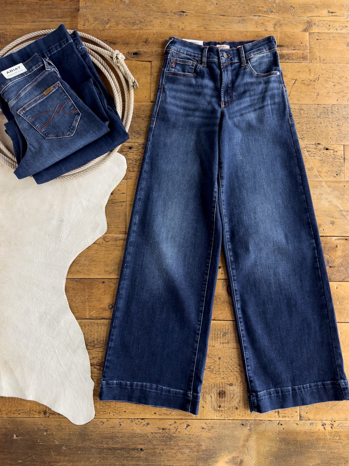 A pair of Kemp High Rise Wide Leg Jeans by Savannah Sevens western life{&}style lies flat on a wooden surface beside a stack of folded jeans, a coiled rope, and a white rug.