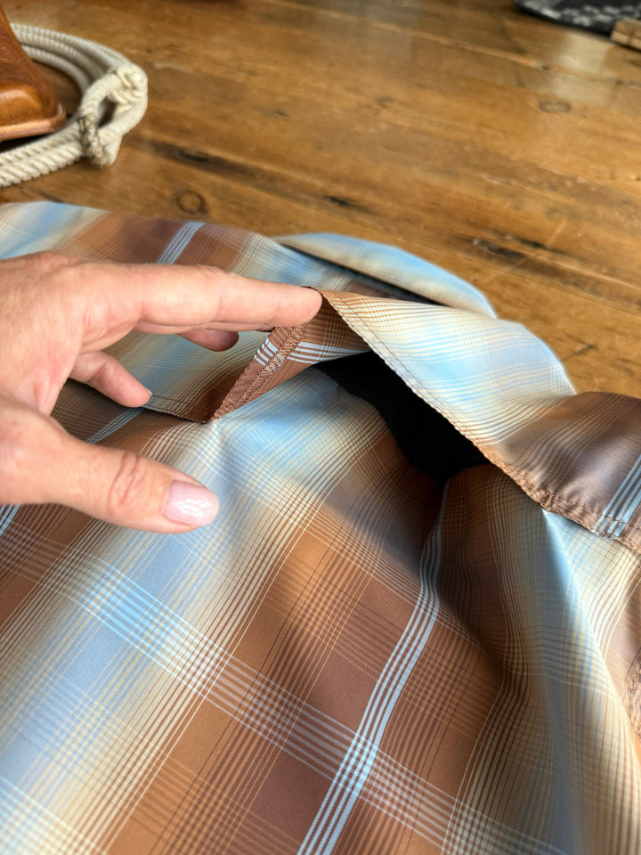 A hand lifts the inner lining of a brown and blue Kent Wrangler Men's Plaid Pearl Snap jacket from Savannah Sevens western life{&}style, revealing a hidden pocket. The shirt rests on a wooden surface with a coiled rope visible in the background.