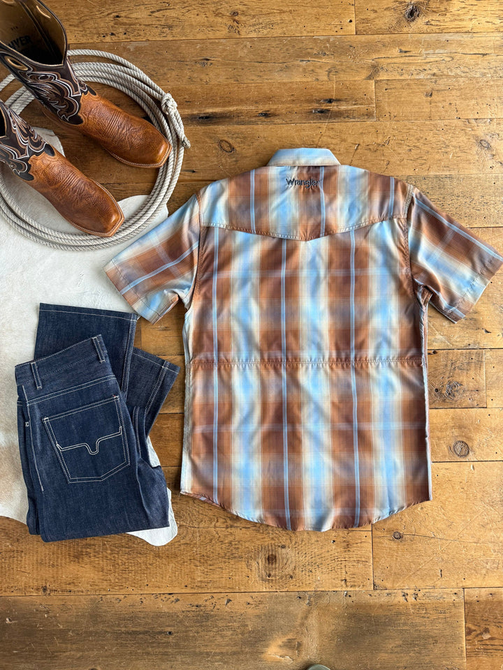 The Kent Wrangler Men's Plaid Pearl Snap by Savannah Sevens western life{&}style, paired with dark blue jeans and cowboy boots, is arranged on a rustic wooden floor to evoke classic Western cowboy style. This short-sleeve shirt delivers moisture-wicking comfort and sun protection, making it ideal for days spent outdoors.