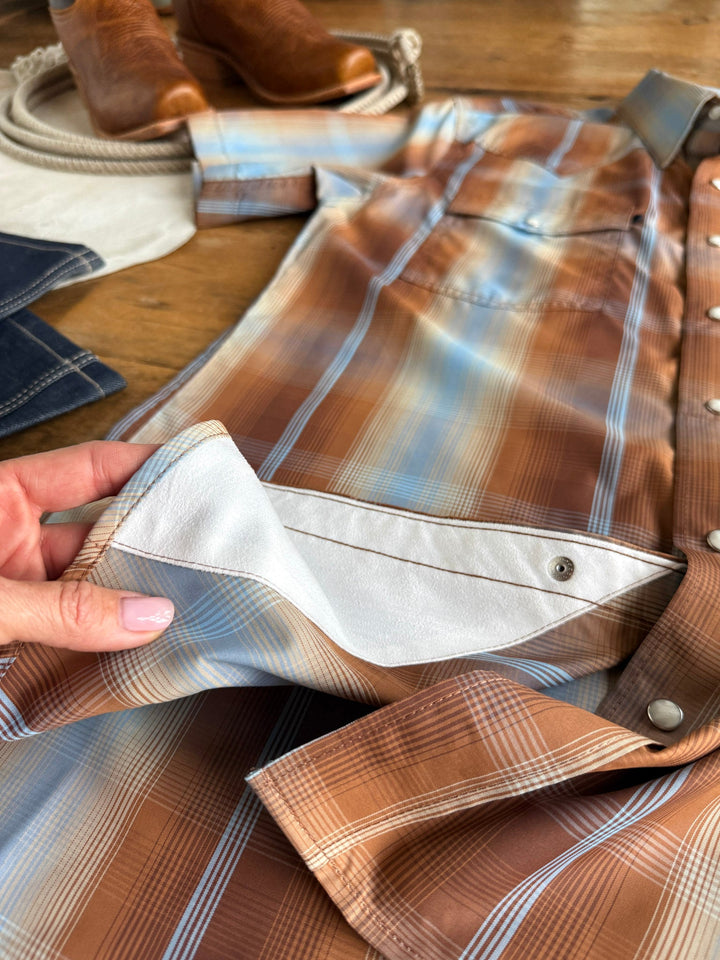 A hand holds open the snap button cuff of the Kent Wrangler Men's Plaid Pearl Snap shirt by Savannah Sevens western life{&}style in brown and blue plaid, revealing the inner white lining. Nearby are brown boots, a coiled rope, and denim jeans set on a wooden surface.