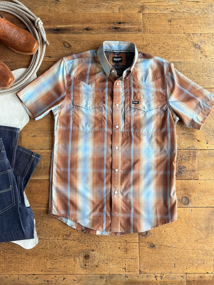 The Kent Wrangler Men's Plaid Pearl Snap by Savannah Sevens western life{&}style—a short-sleeved, pearl snap shirt in light brown, blue, and white plaid—is laid flat on a wooden surface with jeans and cowboy boots nearby. Designed for all terrain adventures, this shirt provides sun protection and moisture-wicking properties for superior comfort wherever you go.