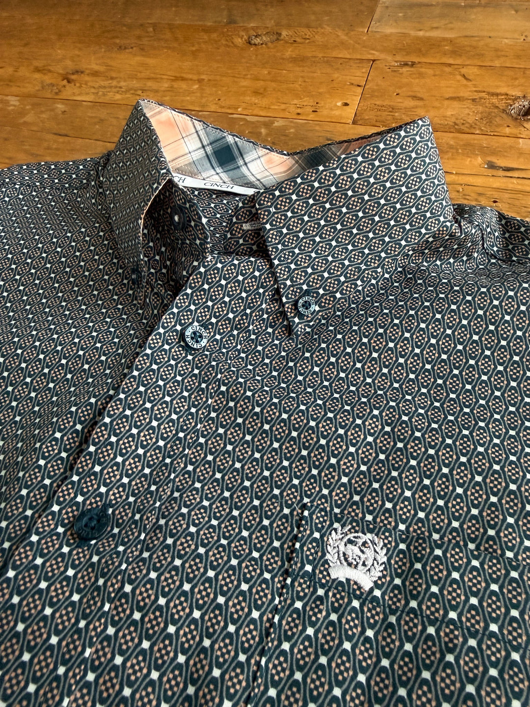 The Kenton Cinch Men’s Southwest Button Up from Savannah Sevens western life{&}style, featuring a striking geometric design, lies on a wooden surface. Its open collar reveals a contrasting plaid lining inside, accentuating its modern Cinch fit.