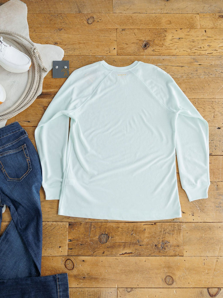 The Kersey Ariat Long Sleeve Top by Savannah Sevens western life{&}style, featuring cooling technology for comfort, is laid flat on a wooden floor beside blue jeans, white shoes, and a gray hanger.