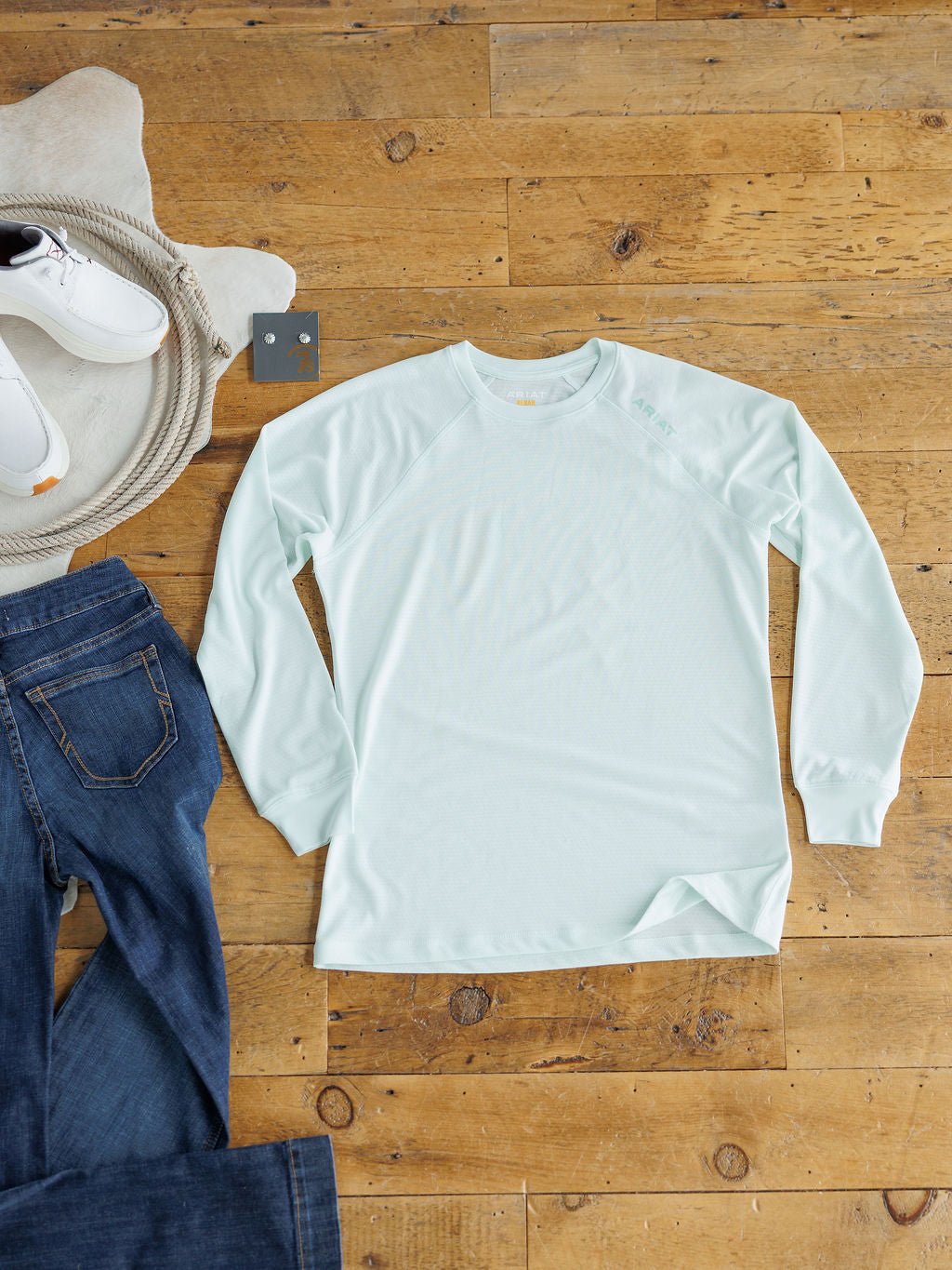 The Kersey Ariat Long Sleeve Top by Savannah Sevens western life{&}style in seafoam mesh with cooling technology is laid flat on a wooden floor beside dark blue jeans, white sneakers, and a round rug.