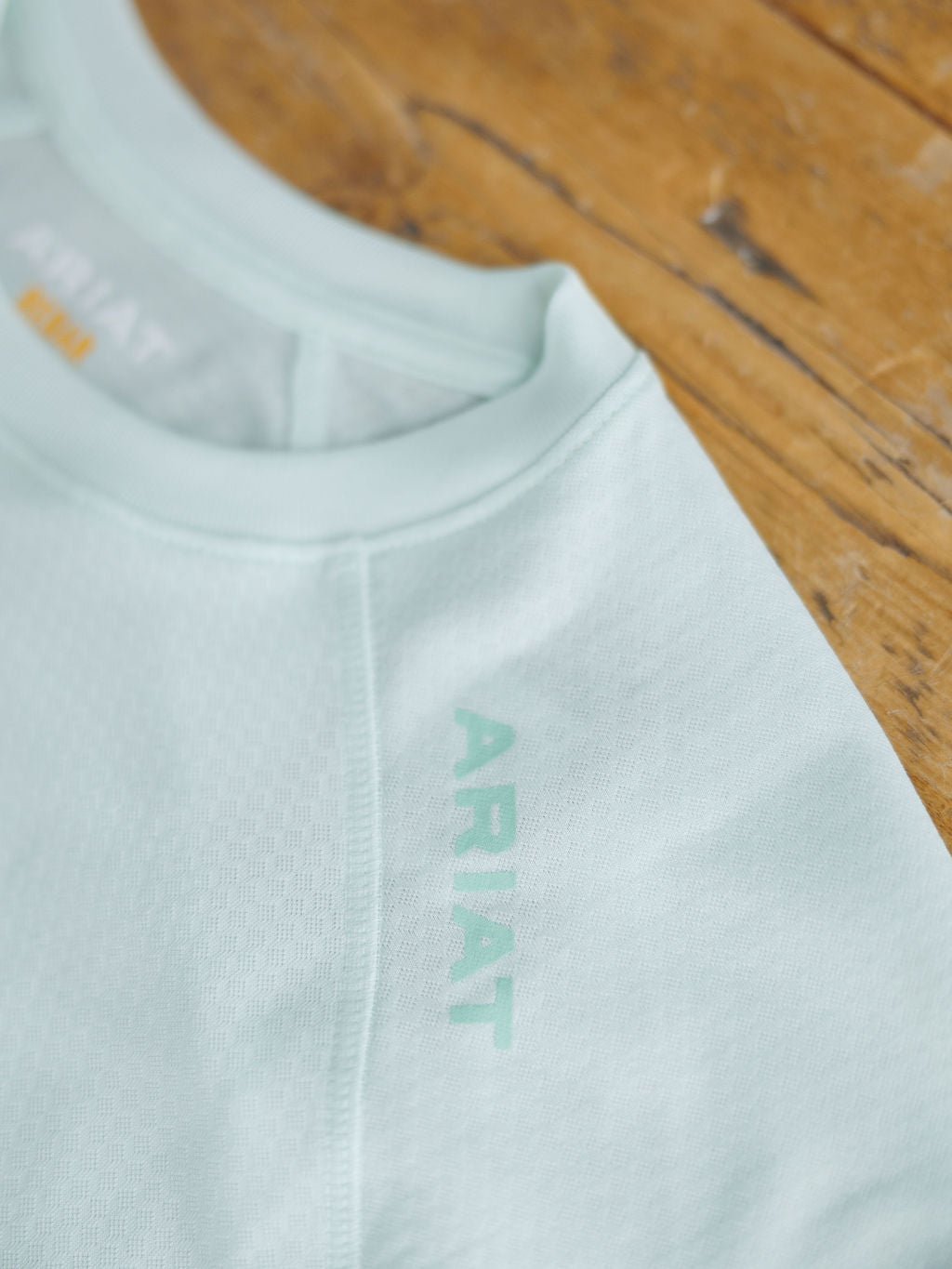 A close-up of the Kersey Ariat Long Sleeve Top by Savannah Sevens western life{&}style in light blue, featuring cooling technology and odor control fabric, with the brand name on the shoulder, displayed on a wooden surface.