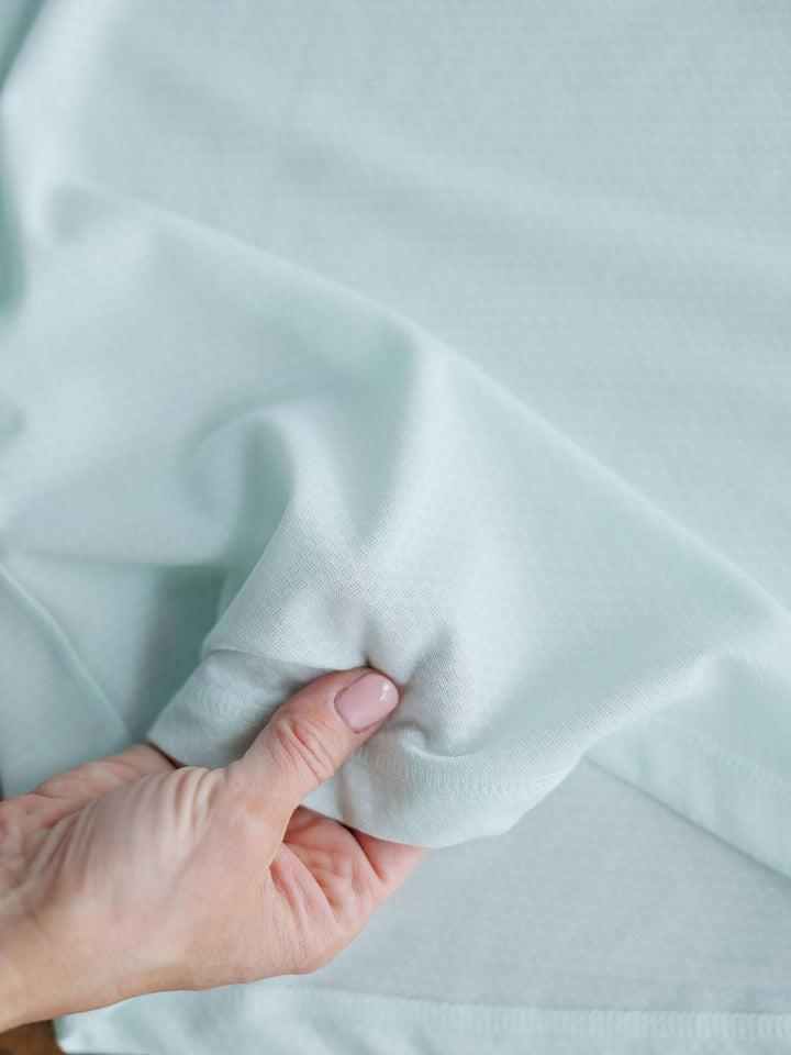 A hand gently lifts the soft, flexible sleeve of the Kersey Ariat Long Sleeve Top by Savannah Sevens western life{&}style, showcasing its light blue, textured fabric.