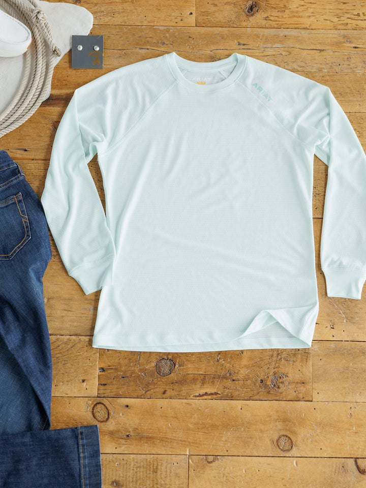 The Kersey Ariat Long Sleeve Top by Savannah Sevens western life{&}style in seafoam mesh with cooling technology is laid flat on a wooden floor next to blue jeans, a tan hat, and a small gray square object.