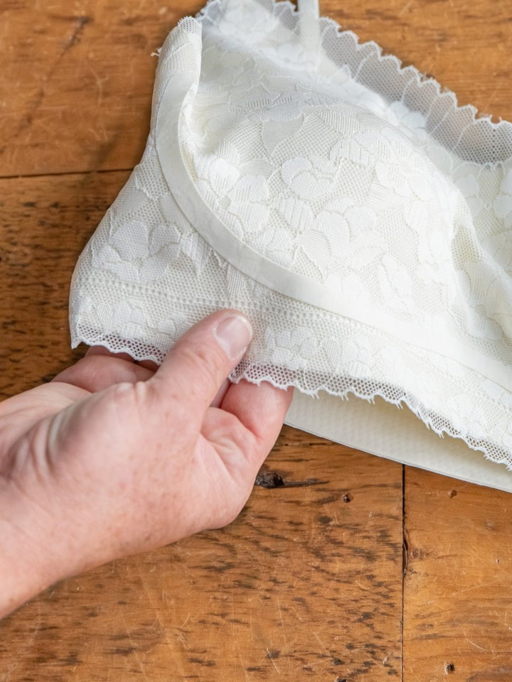 A hand holds the Kersey Bralette by Savannah Sevens western life{&}style against a wooden surface, showcasing its delicate white floral lace pattern, adjustable straps, and the detailed edge of the bra cup.
