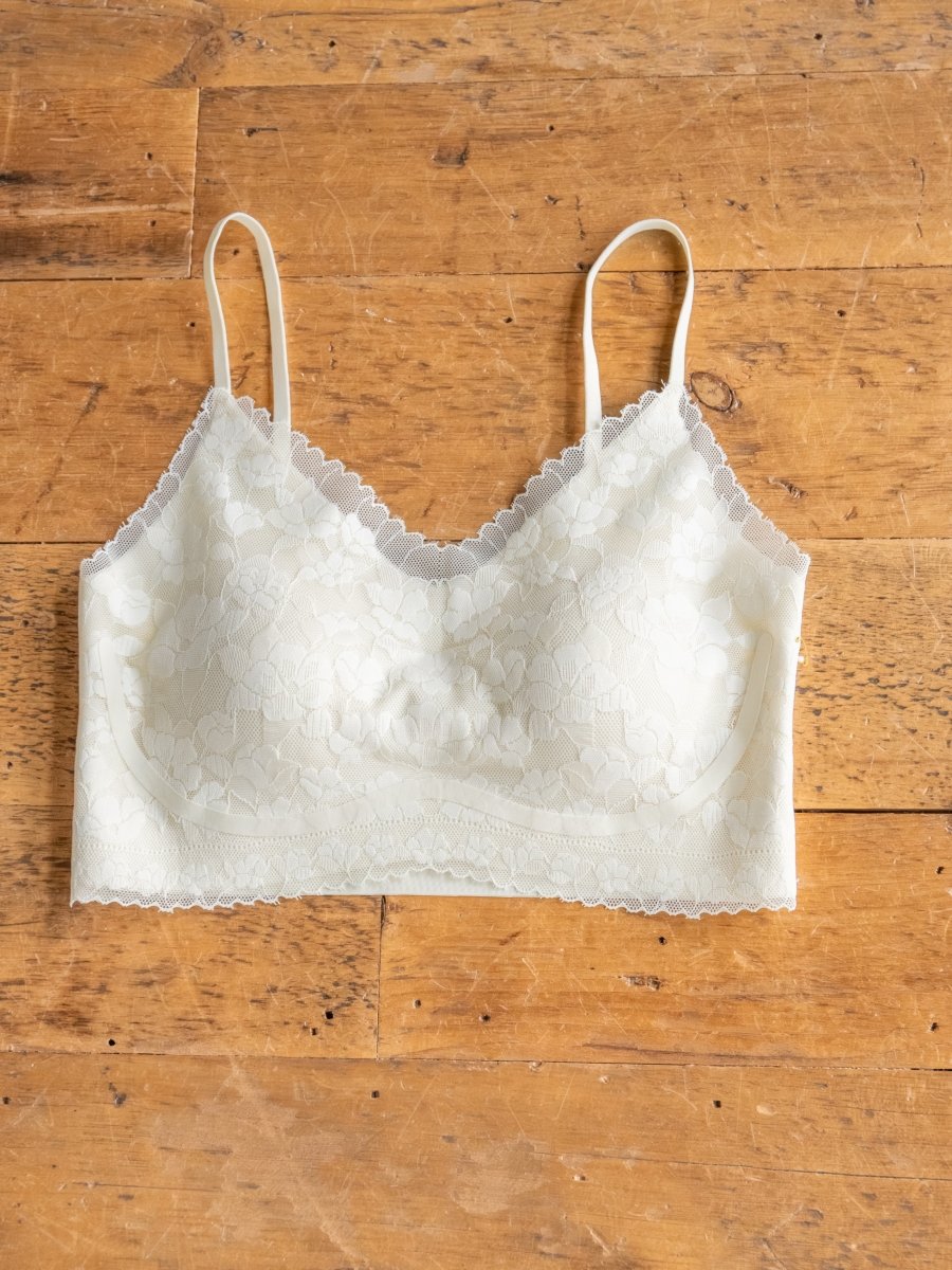 The Kersey Bralette by Savannah Sevens western life{&}style features cream-colored lace, adjustable straps, and scalloped edges, shown laid flat on a wooden surface.