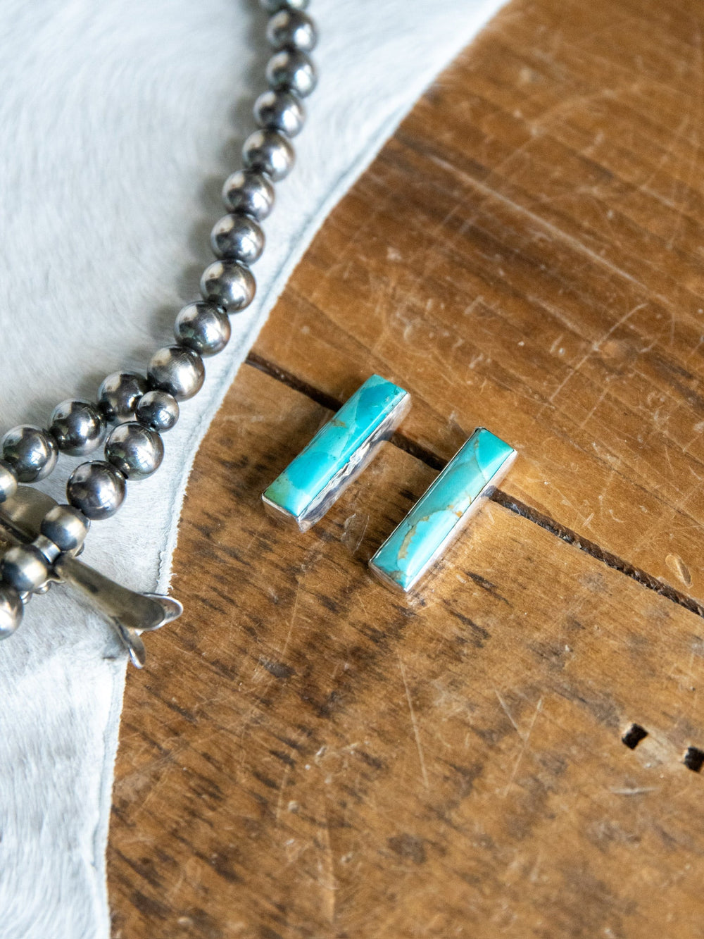 Two rectangular Kewanee Turquoise Bar Earrings by Savannah Sevens western life{&}style, set in sterling silver, rest on a wooden surface beside a beaded silver necklace and a piece of white hide.