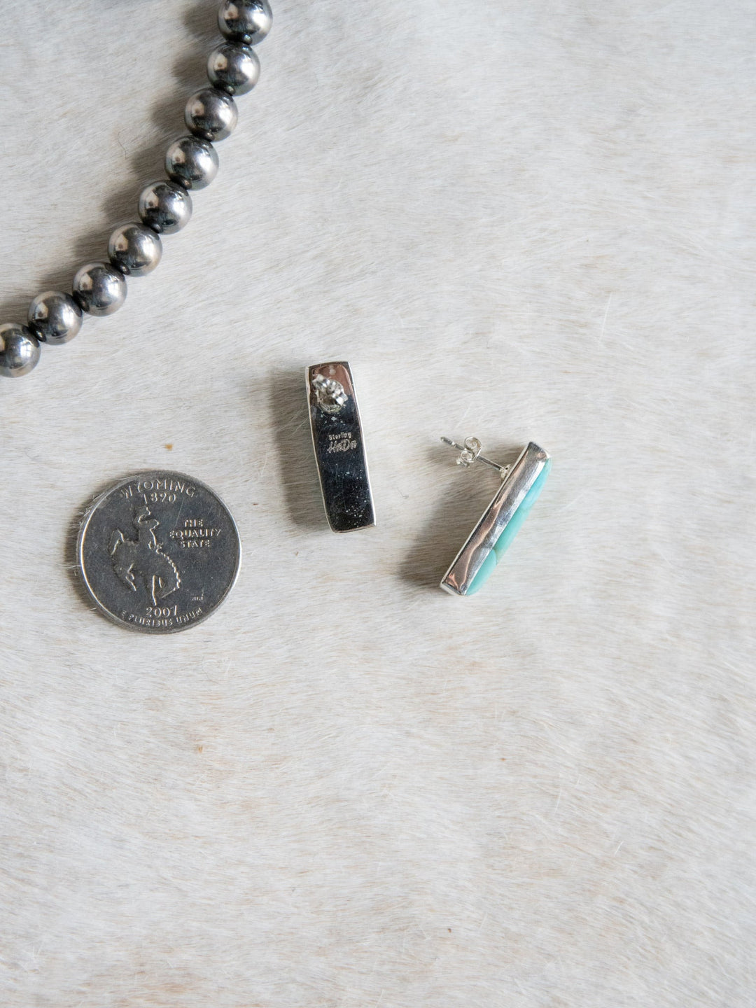 A pair of Kewanee Turquoise Bar Earrings by Savannah Sevens western life{&}style, featuring one earring with a striking turquoise inlay side, is shown next to a 2017 quarter and a silver beaded necklace, all placed on a light textured surface.