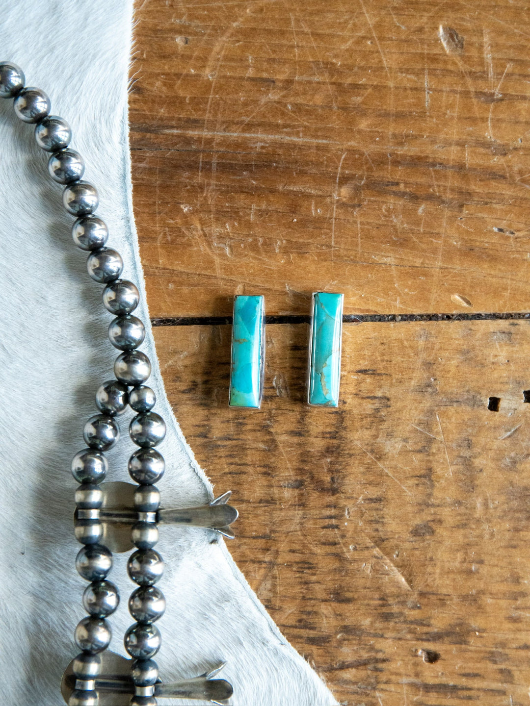 Two rectangular Kewanee Turquoise Bar Earrings by Savannah Sevens western life{&}style rest on a wooden surface next to a string of round sterling silver beads, partially on a piece of white fur.