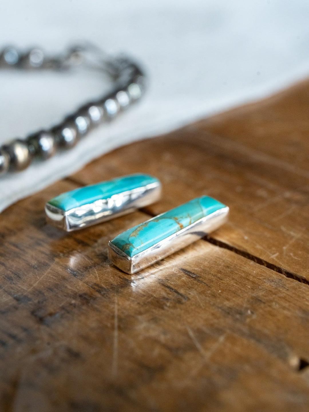 Two Kewanee Turquoise Bar Earrings by Savannah Sevens western life{&}style, crafted in sterling silver and showcasing vibrant turquoise stones, rest on a rustic wooden surface while a beaded silver necklace lingers softly out of focus in the background.