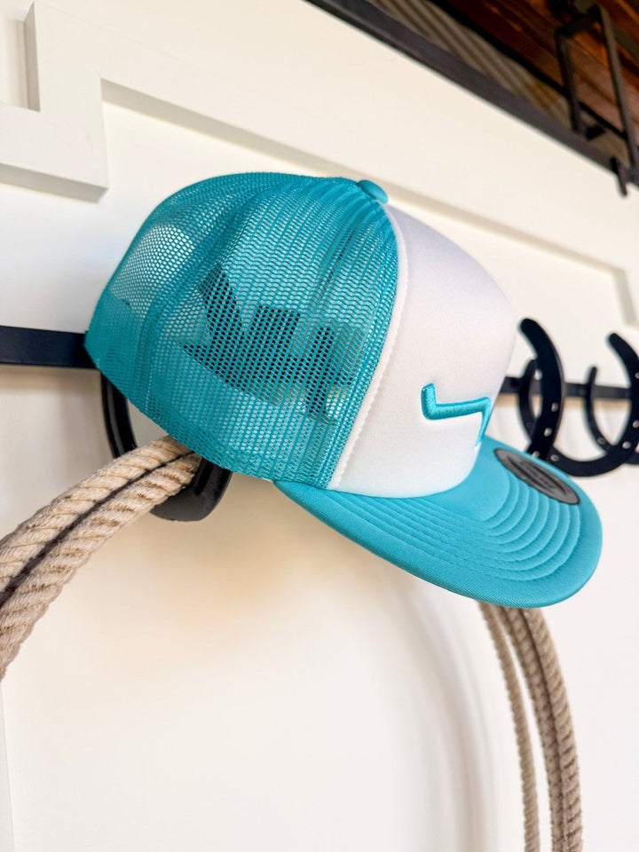 The Kimes Durant Cap from Savannah Sevens western life{&}style features turquoise and white colors, turquoise embroidery, a wave logo, and an adjustable snap back. It’s displayed on a coiled rope with black horseshoes in the background.