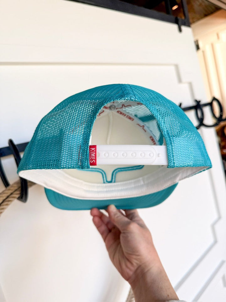 A hand holds the Kimes Durant Cap by Savannah Sevens western life{&}style upside down, revealing its adjustable snap back, turquoise embroidery, red KIMES tag inside, and a background with white paneling and black hooks.