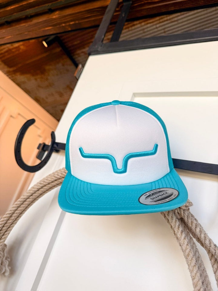 The Kimes Durant Cap by Savannah Sevens western life{&}style features white and teal panels, blue bullhorn embroidery, and an adjustable snapback. It hangs on a black horseshoe hook with a thick rope nearby against a white door.