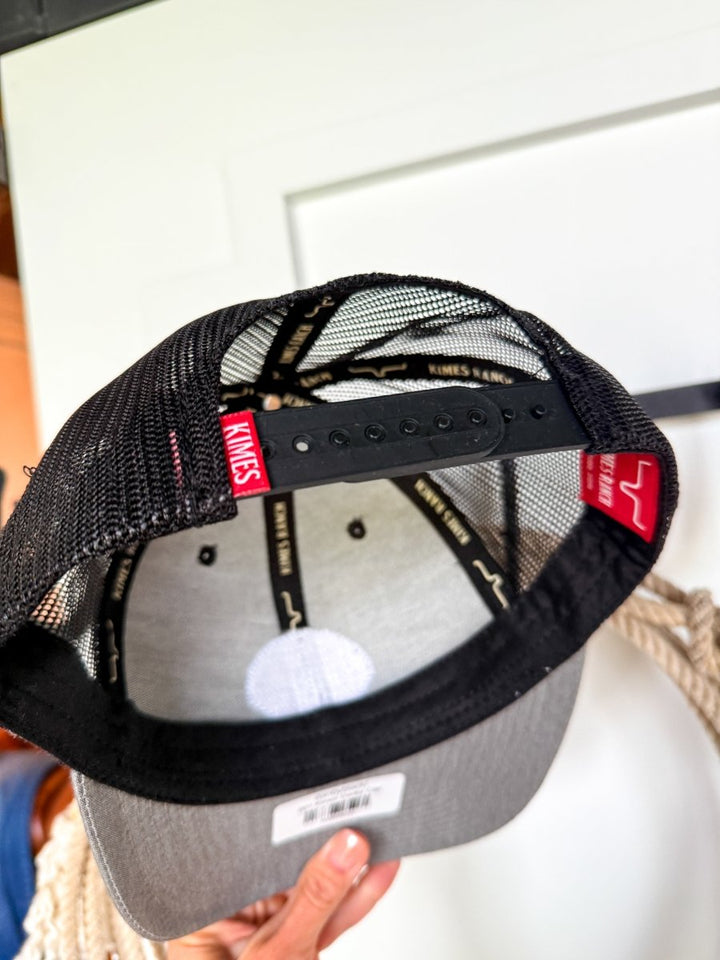 A hand holds a black and gray mesh Kimes Ranch Cap, displaying the inside. The inner sweatband features “Savannah Sevens western life{&}style,” and an adjustable snap back closure is visible at the back.