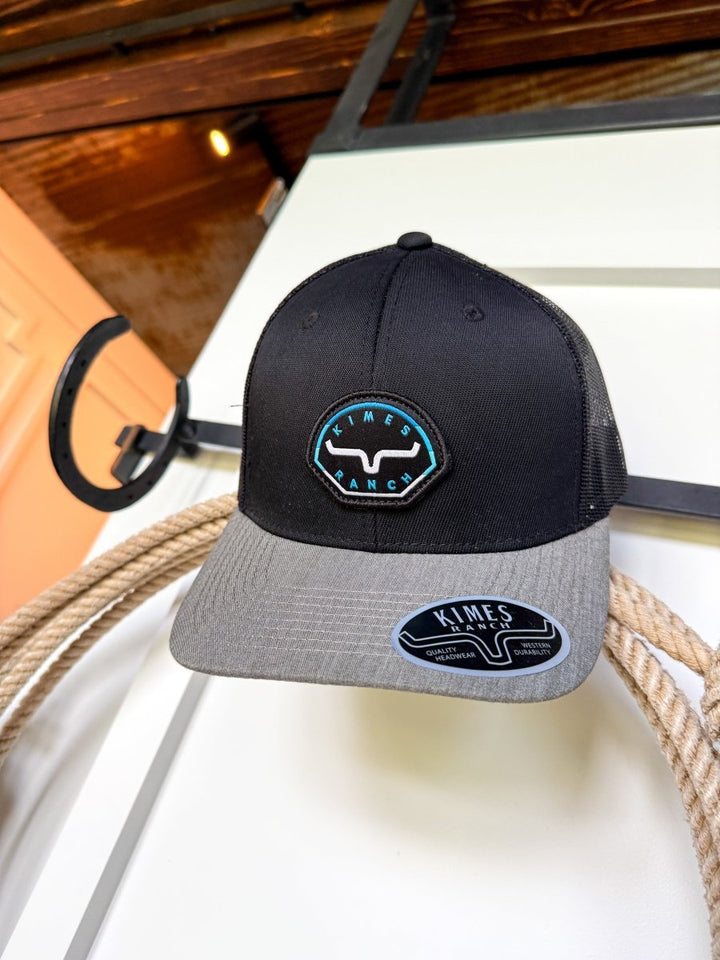 The Kimes Ranch Cap by Savannah Sevens western life{&}style, in black and gray with a bull logo patch, rests on a coiled rope before a white and wood wall with a black horseshoe. This snapback features classic southwest styling.