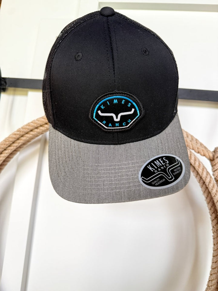 A Kimes Ranch Cap from Savannah Sevens western life{&}style, in black and gray with a blue and white southwest logo patch, hangs on a coiled rope. It features an adjustable snap back and branded sticker on the gray brim.