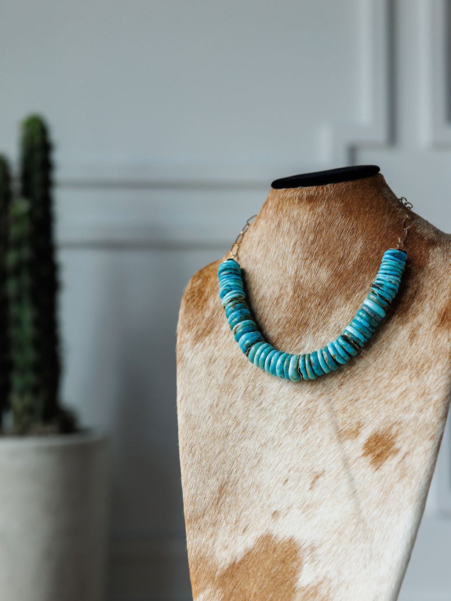 The Kingman Necklace by Savannah Sevens western life{&}style, featuring turquoise, is showcased on a tan and white cowhide mannequin with a potted cactus softly blurred in the white-walled background.