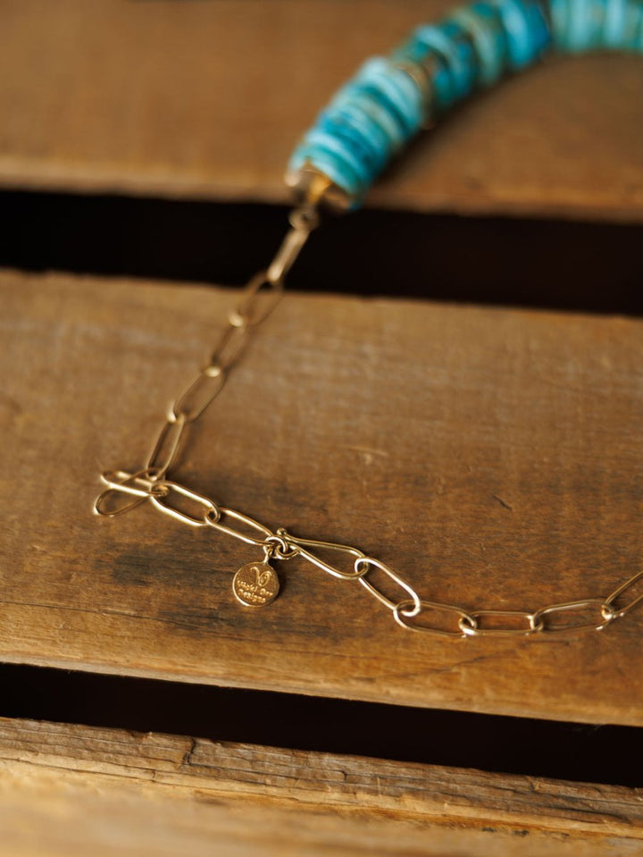 The Kingman Necklace by Savannah Sevens western life{&}style features a gold paperclip chain with turquoise beads and a round charm, shown in close-up on a rustic wooden surface.